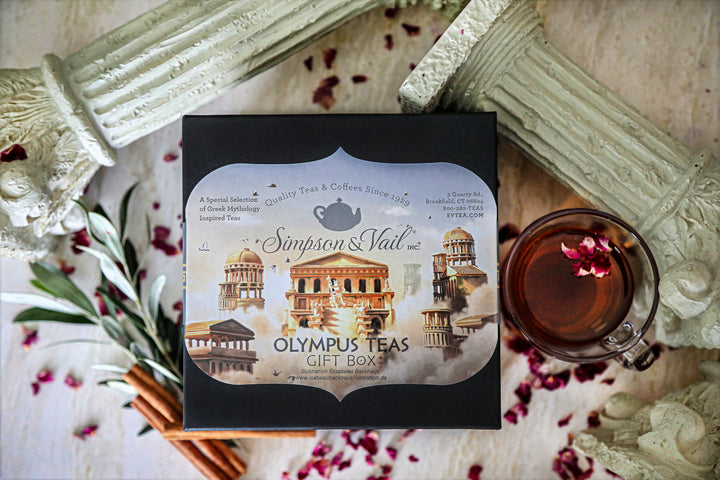 Simpson &amp; Vail Olympus Teas Gift Box in black packaging, styled in a flat-lay with a glass of tea, cinnamon sticks, rose petals, and classical column props on a light wooden surface.