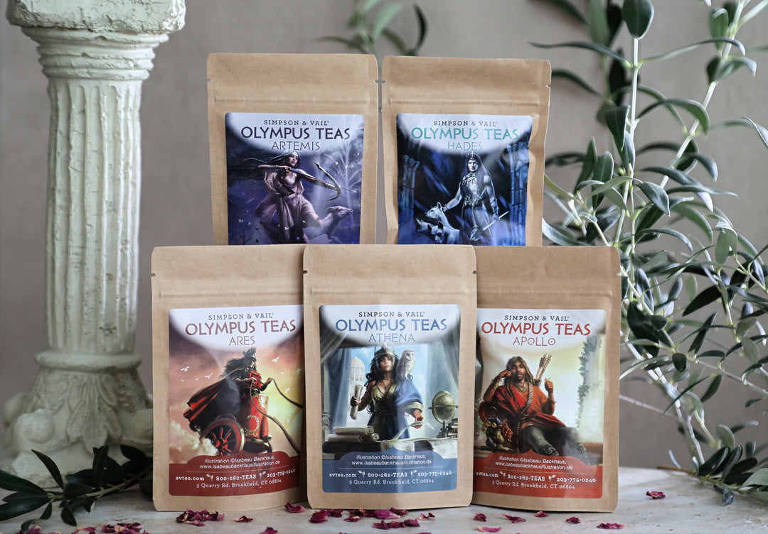 Five stand-up pouches of Olympus Teas fantasy tea blends arranged on a tabletop with decorative stone and greenery in the background. The kraft paper bags feature colorful illustrated labels for various character teas.