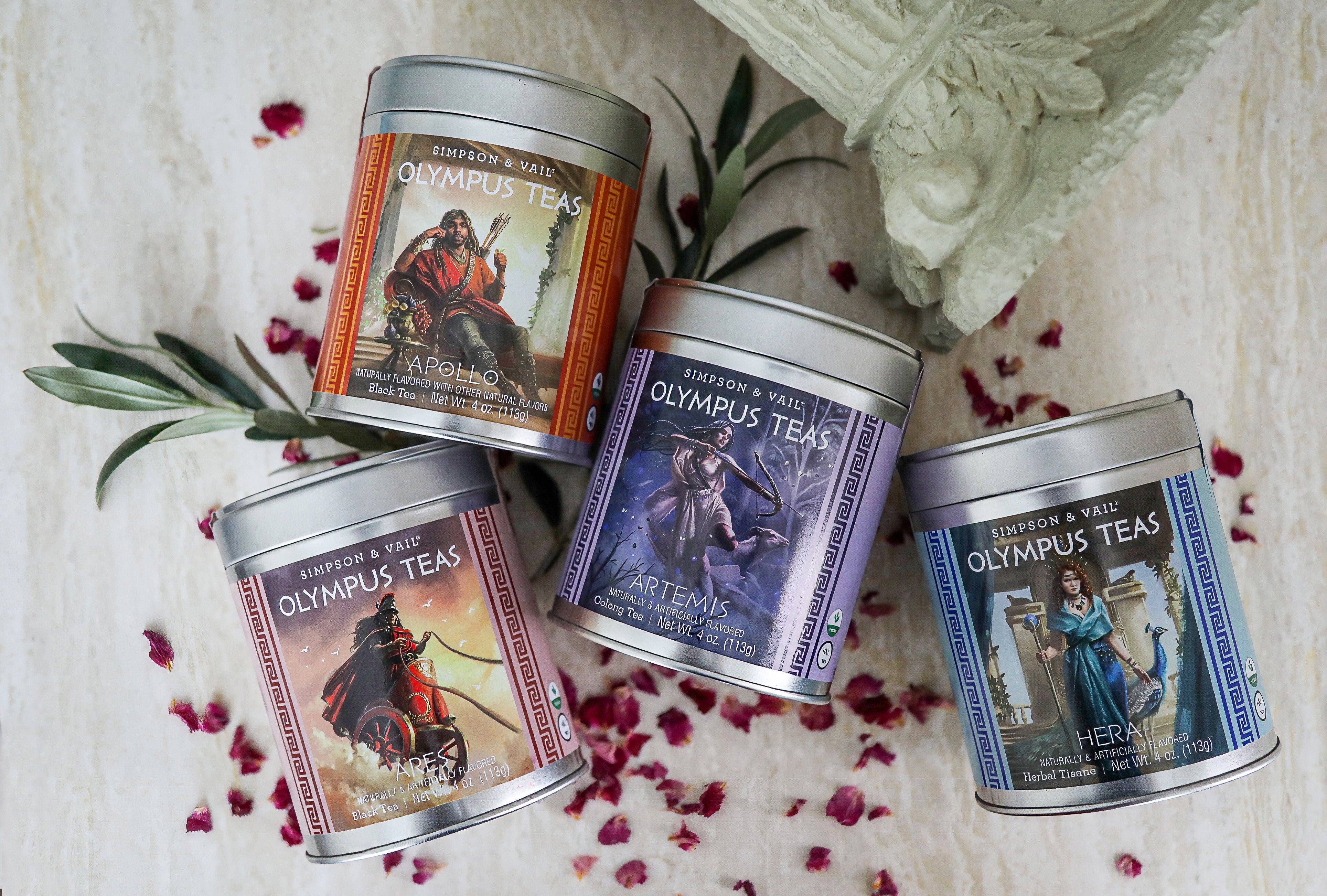 Four metal tins of Olympus Teas arranged on a light stone surface with scattered rose petals and greenery. The tins feature fantasy-themed labels in orange, purple, pink, and blue, with silver lids. Apollo, Ares, Artemis, Hera