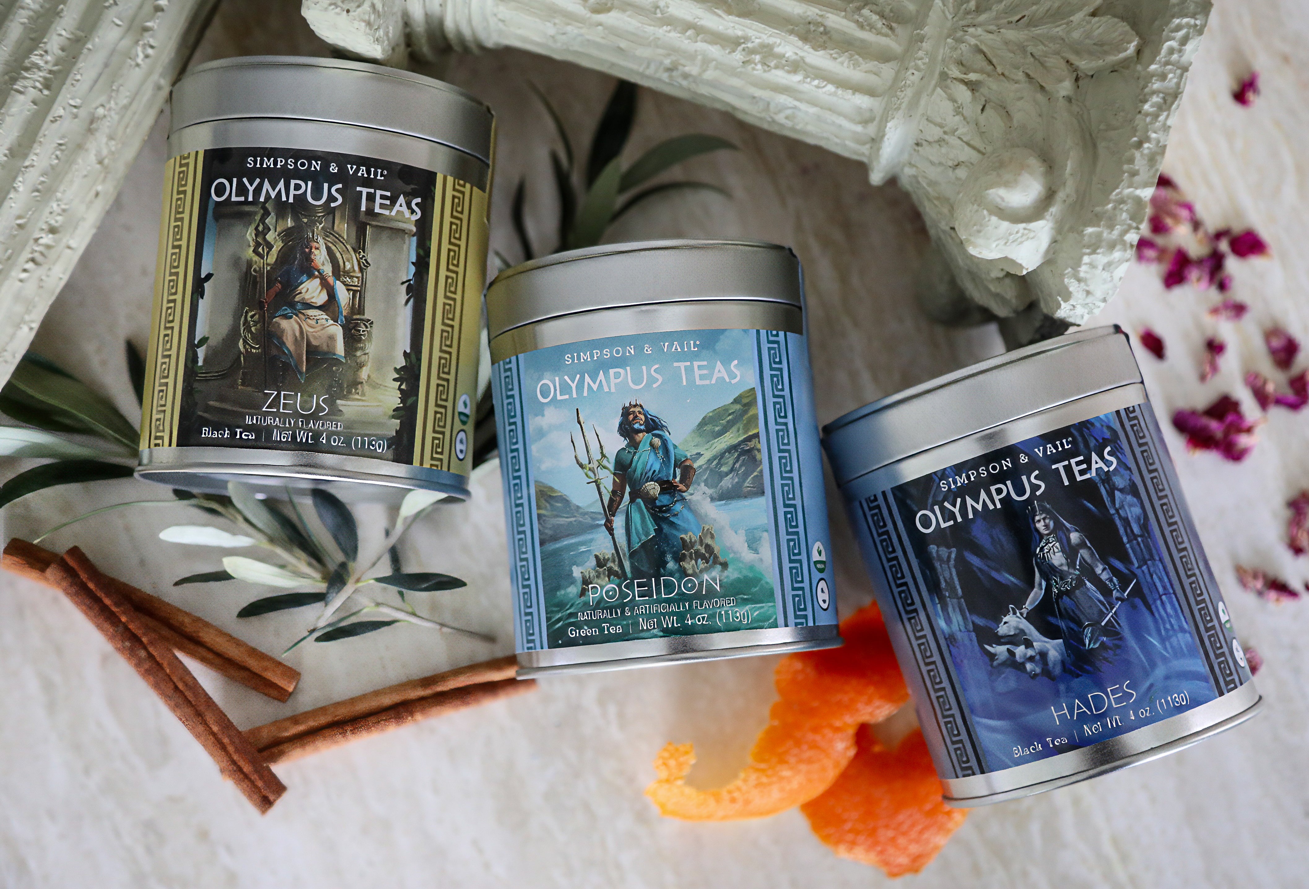 Three round metal tins of Simpson & Tail Olympus Teas arranged on a white surface with decorative props including cinnamon sticks, orange peel, dried flowers, and a white architectural element. The labels show mythological figures and the names Zeus, Poseidon, and Hades.