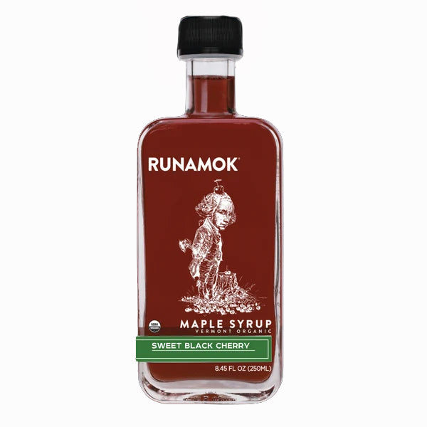 A bottle of Runamok Sweet Black Cherry maple syrup on a plain white background. The bottle is clear glass showing the dark red syrup inside, with a black cap and a label featuring a white illustration of a George Washington next to a chopped down cherry tree with a cherry on his head and a green band labeled &